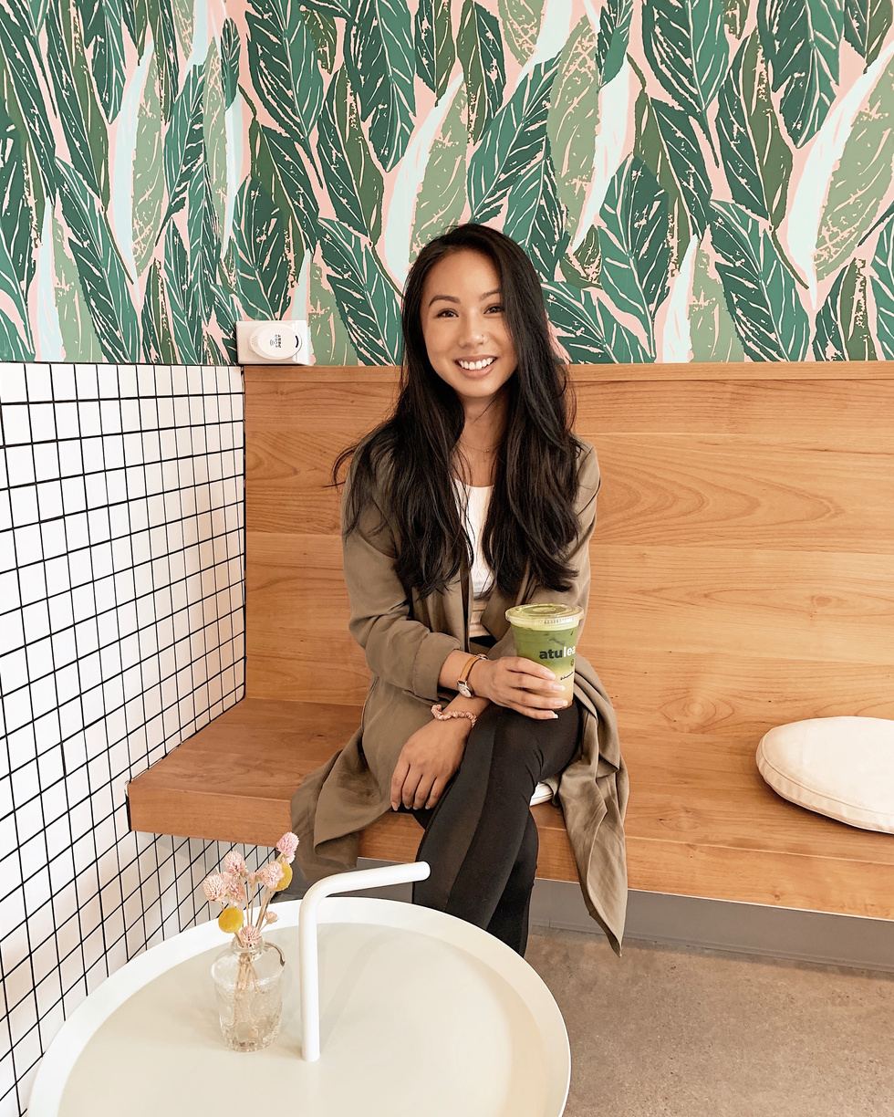 A young woman sitting on a bench smiling holding a matcha latte at a cute cafe in Seattle called Atulea