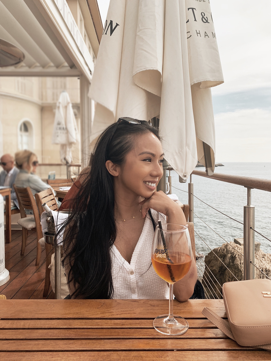 a young woman sitting at a table at an outdoor restaurant in France looking out into the ocean with a glass of Aperol Spritz on her table